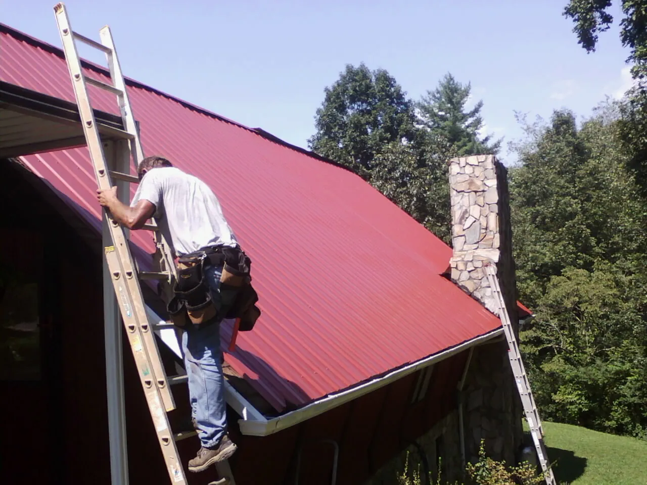Roofer on a ladder beside a red metal roof and stone chimney — Soffit & Fascia Repair by Roofing Brothers in Burbank